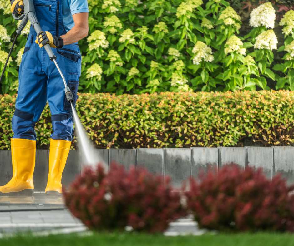 A gardener in blue overalls and yellow boots pressure washing a walkway, surrounded by lush greenery and well-kept bushes, demonstrating effective ways to keep a garden clean.
